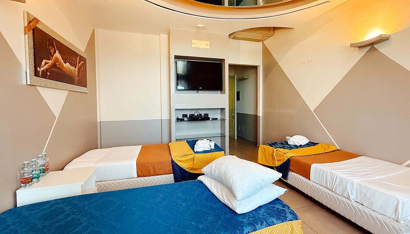 Hotel room with three single beds (blue, orange, and white linens), towels on two beds, and a TV mounted on the wall; modern geometric wall design in beige and gray.